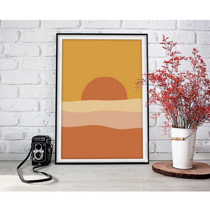 May include: A framed print of a minimalist sunset landscape with a yellow, orange, and brown color scheme. The sun is a large orange circle above a series of wavy lines representing the horizon.
