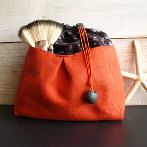 May include: A small orange linen drawstring bag with a black and white floral print lining. The bag has a black tassel with a round, dark gray bead.