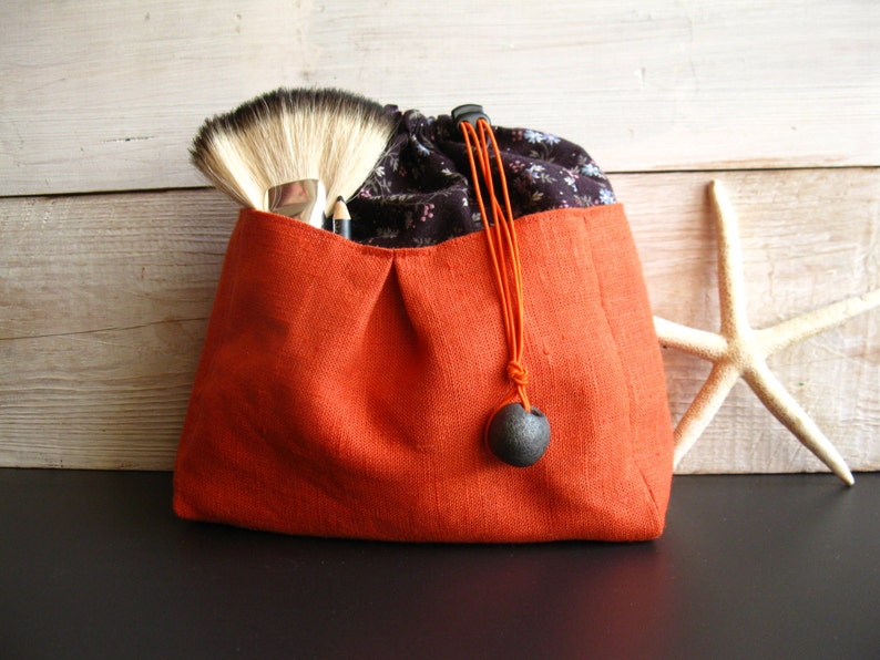 May include: A small orange linen drawstring bag with a black and white floral print lining. The bag has a black tassel with a round, dark gray bead.