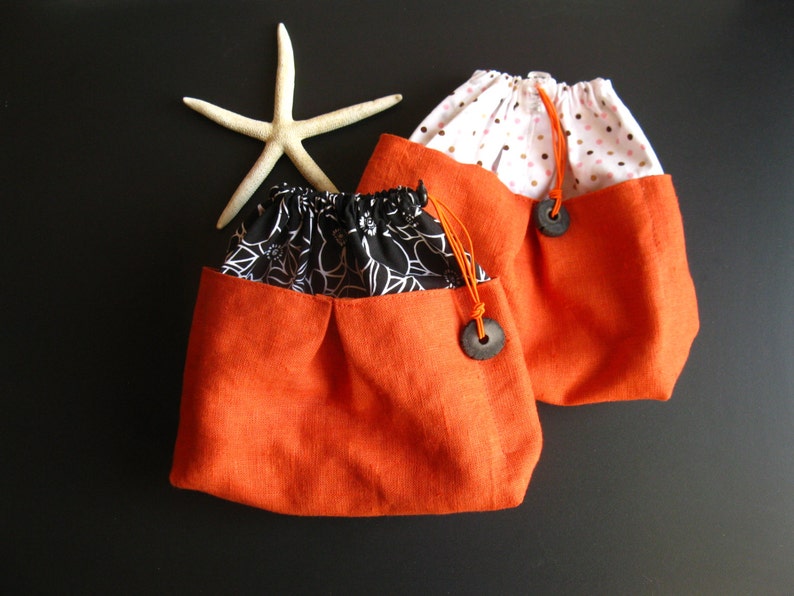 May include: Two orange drawstring bags with black and white floral fabric lining. One bag has a white background with pink polka dots.