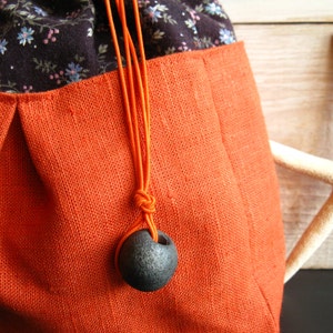 May include: A drawstring bag made of orange linen with a black floral print lining. The bag has a black cord with a dark gray ceramic bead.