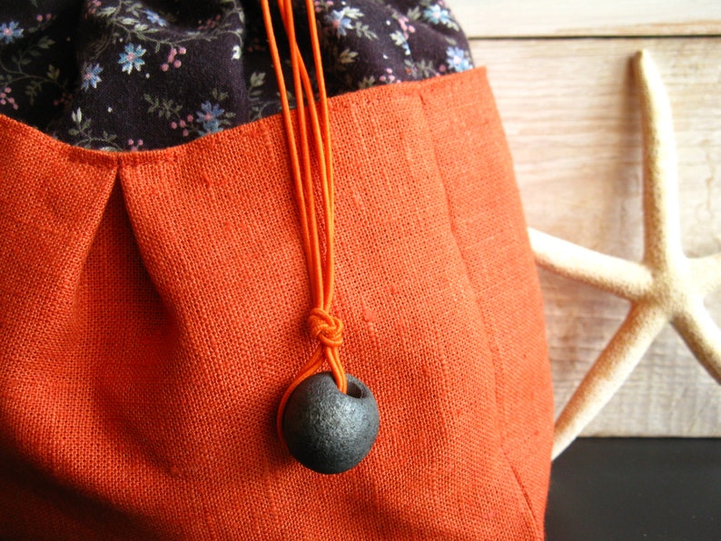 May include: A drawstring bag made of orange linen with a black floral print lining. The bag has a black cord with a dark gray ceramic bead.