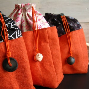 May include: Three drawstring bags with orange fabric bottoms and different patterned tops. The bags have black and white floral patterns, black and white geometric patterns, and pink floral patterns. Each bag has a black or white decorative accent on the drawstring.