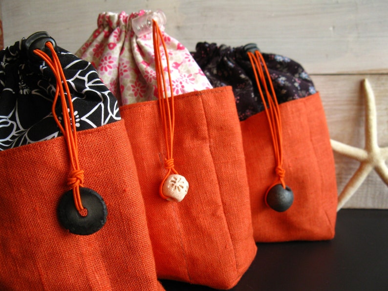 May include: Three drawstring bags with orange fabric bottoms and different patterned tops. The bags have black and white floral patterns, black and white geometric patterns, and pink floral patterns. Each bag has a black or white decorative accent on the drawstring.