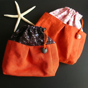 May include: Two orange fabric drawstring bags with different patterned fabric lining. The bags have a small, round, dark brown button on the drawstring.
