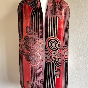 May include: A long, Art Deco-style scarf in shades of red, burgundy, and gold. The scarf features vertical stripes and circular patterns. The fabric appears to be velvet or a similar plush material, draped over a mannequin.