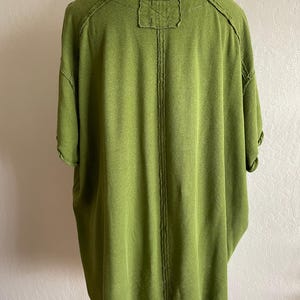May include: A short-sleeved, olive green shirt with a relaxed fit. The back of the shirt features a vertical seam down the center and a small, rectangular patch at the neckline. The fabric has a textured appearance.
