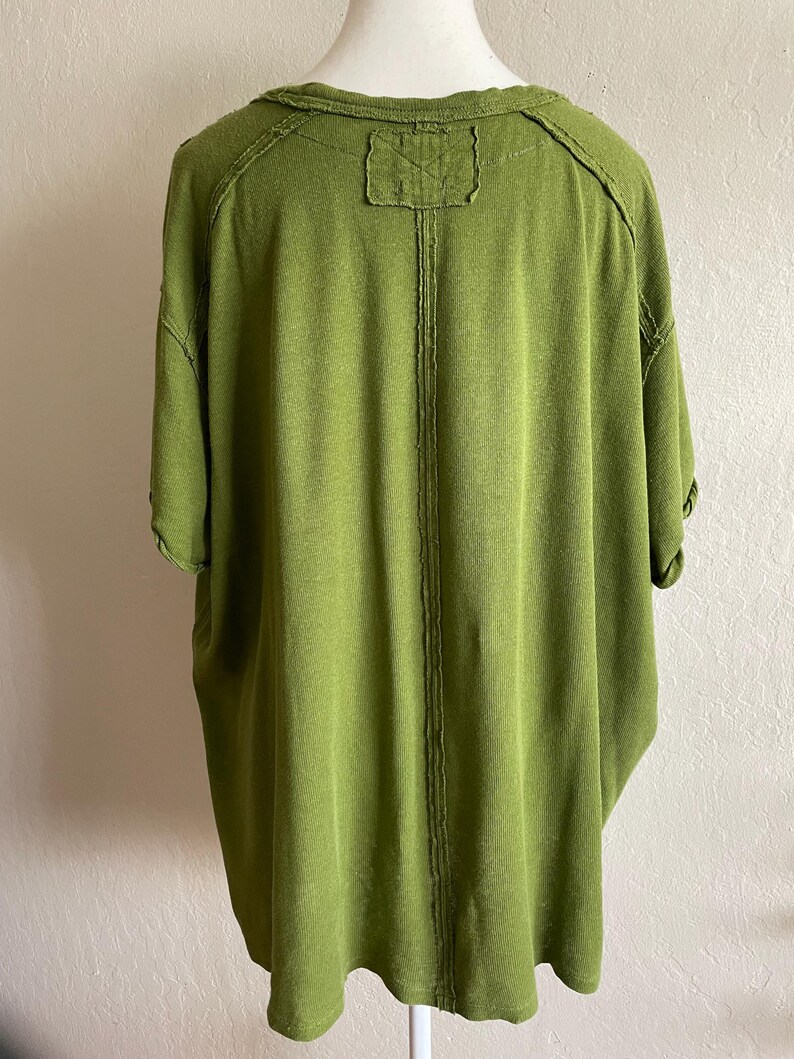 May include: A short-sleeved, olive green shirt with a relaxed fit. The back of the shirt features a vertical seam down the center and a small, rectangular patch at the neckline. The fabric has a textured appearance.