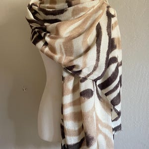May include: A beige, brown, and white patterned scarf draped over a mannequin. The scarf features a zebra stripe design with fringed edges. The scarf is made of a soft, lightweight material.