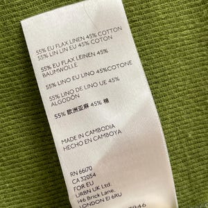 May include: A white clothing tag with text in multiple languages, including English, French, and Spanish, detailing the fabric composition as 55% flax linen and 45% cotton. The tag also indicates the garment was made in Cambodia.