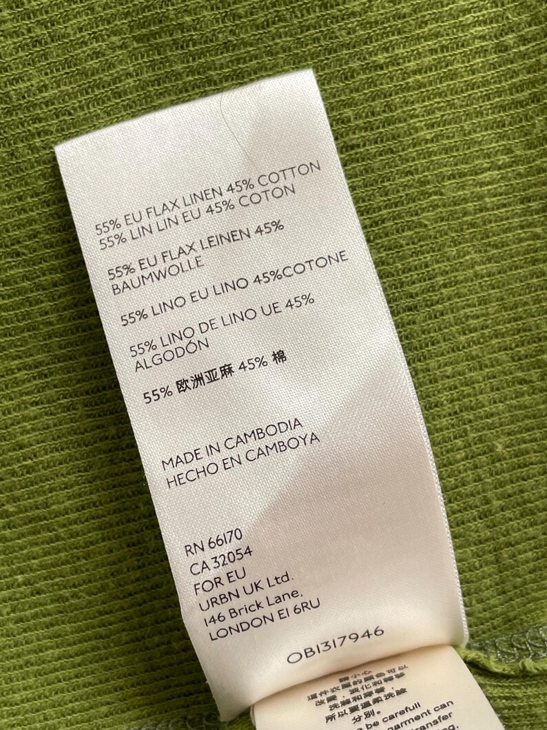 May include: A white clothing tag with text in multiple languages, including English, French, and Spanish, detailing the fabric composition as 55% flax linen and 45% cotton. The tag also indicates the garment was made in Cambodia.