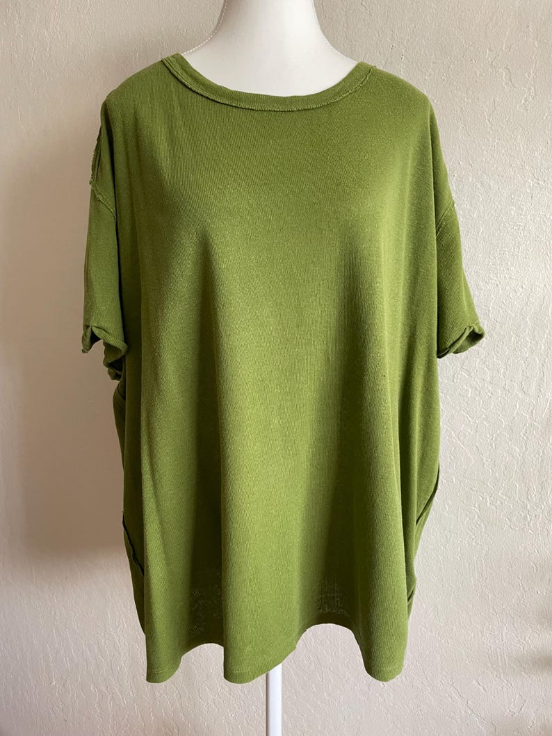 May include: Olive green short-sleeved top with a round neckline. The shirt has a loose, oversized fit and a slightly textured appearance. The sleeves are cuffed, and the fabric appears to be a soft knit material. The top is displayed on a mannequin.