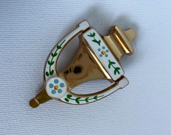 May include: A gold-toned brooch with a decorative design. The brooch features white enamel accents with blue and red floral details and green leaf patterns. The brooch has a horseshoe shape.