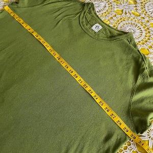 May include: Olive green t-shirt with a crew neck. A yellow measuring tape is laid across the shirt, showing measurements up to 32 inches. The shirt has a small tag on the neckline. The fabric appears to be soft and slightly textured.