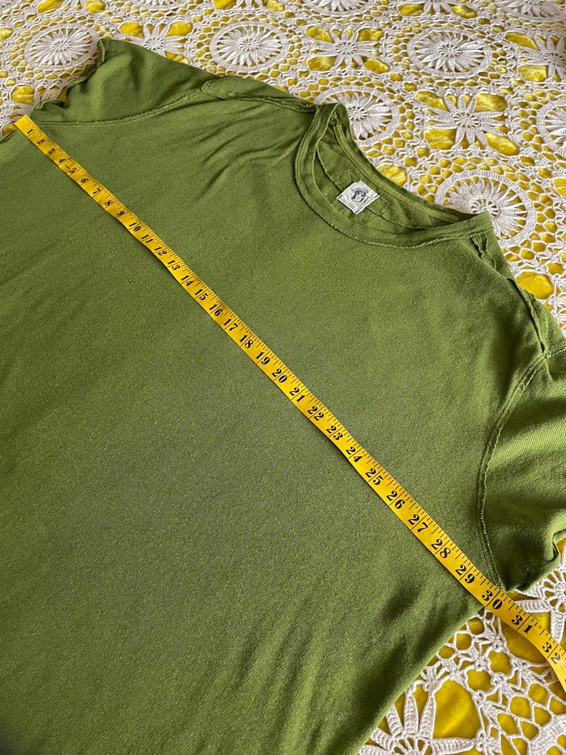 May include: Olive green t-shirt with a crew neck. A yellow measuring tape is laid across the shirt, showing measurements up to 32 inches. The shirt has a small tag on the neckline. The fabric appears to be soft and slightly textured.