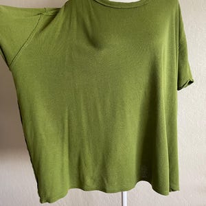 May include: Olive green short-sleeve t-shirt with a round neckline. The shirt has a relaxed fit and is displayed on a white mannequin. The fabric appears soft and drapes loosely. The color is a muted, earthy green.