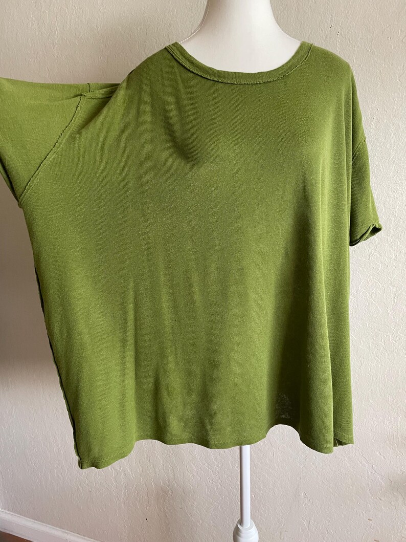 May include: Olive green short-sleeve t-shirt with a round neckline. The shirt has a relaxed fit and is displayed on a white mannequin. The fabric appears soft and drapes loosely. The color is a muted, earthy green.