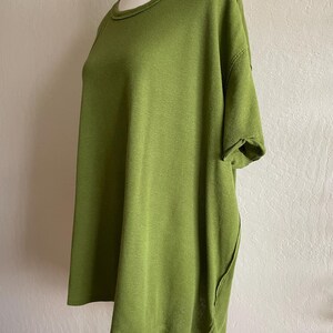 May include: A short-sleeved, olive green top with a rounded neckline. The fabric appears to be a soft knit. The top has a relaxed fit and a unique side seam detail. The garment is displayed on a mannequin.