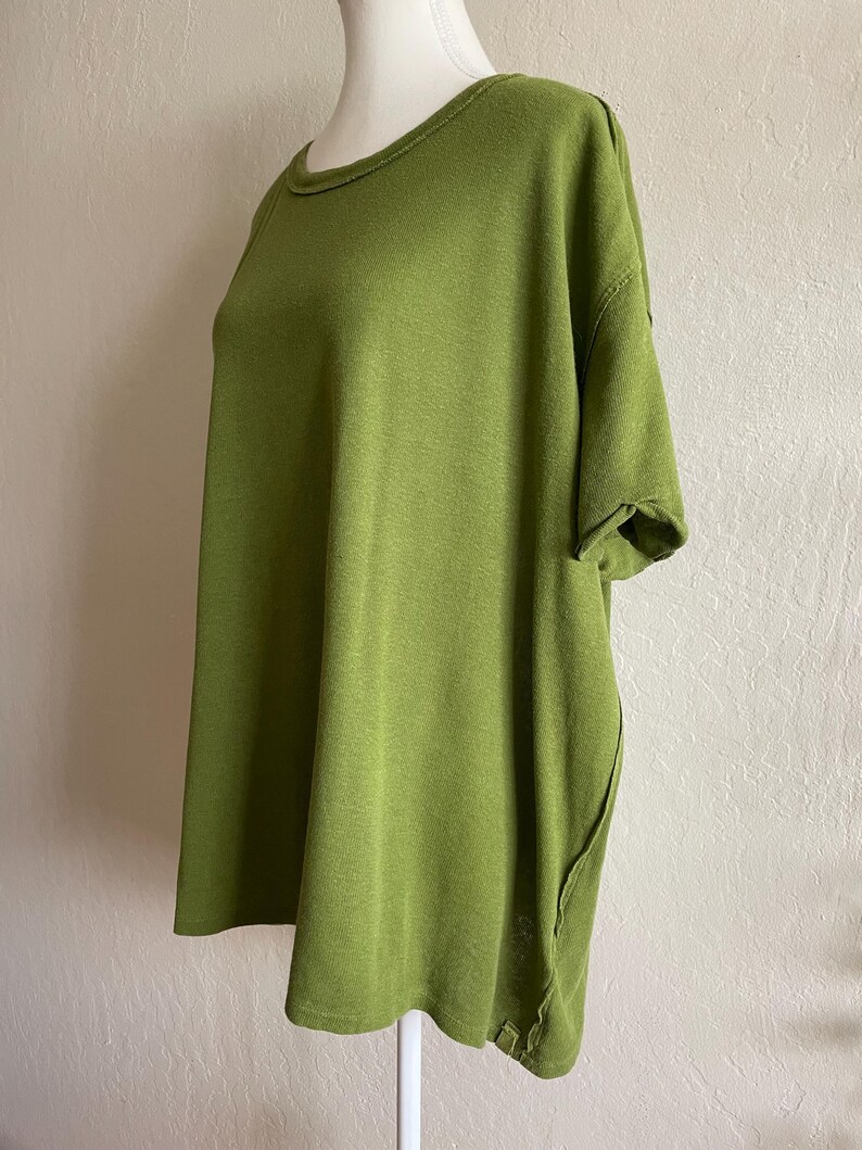 May include: A short-sleeved, olive green top with a rounded neckline. The fabric appears to be a soft knit. The top has a relaxed fit and a unique side seam detail. The garment is displayed on a mannequin.