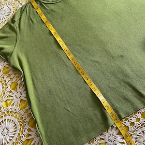 May include: Olive green t-shirt with a round neckline. A yellow measuring tape is laid across the shirt, showing a length of approximately 33 inches. The shirt is on a yellow lace surface.