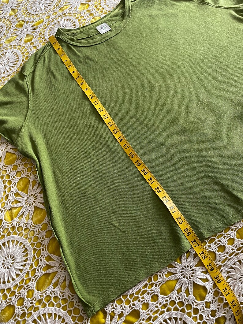 May include: Olive green t-shirt with a round neckline. A yellow measuring tape is laid across the shirt, showing a length of approximately 33 inches. The shirt is on a yellow lace surface.