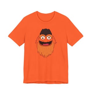 Puede incluir: Camiseta naranja con un personaje de dibujos animados. El personaje tiene un sombrero marrón, pelaje naranja y una boca abierta. La camiseta es de color naranja liso.