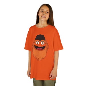 Könnte beinhalten: Orangefarbenes T-Shirt mit einem Cartoon-Charakter. Der Charakter hat einen schwarzen Hut, orangefarbenes Fell und ein breites Lächeln. Das Gesicht des Charakters mit großen Augen und einem fröhlichen Ausdruck ist das zentrale Designelement.