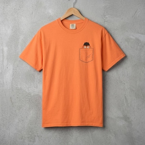 Könnte beinhalten: Orangefarbenes T-Shirt mit kurzen Ärmeln und einem Taschen-Design. Die Tasche zeigt ein herauslugendes Cartoon-Gesicht. Das T-Shirt hängt an einem Holzbügel vor grauem Hintergrund. Das T-Shirt ist aus einem weichen Material.