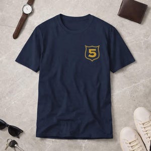Può includere: T-shirt blu navy con un design a scudo dorato con il numero 5. La maglietta a maniche corte ha un'emblema dorato sul petto sinistro. La maglietta è realizzata in materiale morbido ed è esposta su una superficie grigia.