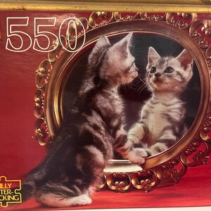 May include: A 550 piece jigsaw puzzle featuring a close-up of two tabby kittens looking at each other in a gold ornate mirror. The puzzle box has a red background with gold accents.