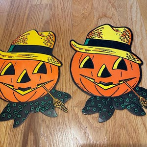 May include: Two vintage Halloween decorations shaped like pumpkins with straw hats and a green and black patterned scarf. The pumpkins have orange faces with black eyes, a nose, and a mouth.