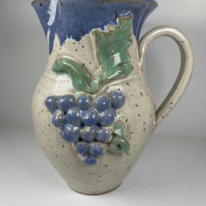 May include: A ceramic pitcher with a blue rim and handle. The pitcher is decorated with a cluster of blue grapes and green leaves. The body of the pitcher is speckled with dark spots.