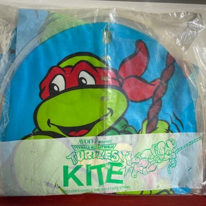 May include: A Teenage Mutant Ninja Turtles kite featuring a green turtle with a red bandana. The kite is packaged in a clear plastic bag with the text "AVON Presents Teenage Mutant Ninja Turtles Kite" and "Includes handle and 300-ft kite string".