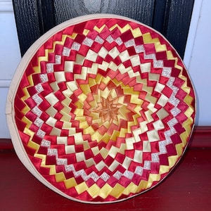 May include: A round, cream-colored pillow with a circular design made of red, yellow, and white ribbon. The ribbon is woven in a spiral pattern, creating a geometric design.