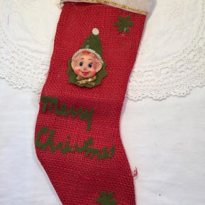 May include: Red burlap Christmas stocking with a white cuff and gold trim. The stocking features a felt elf face and green felt accents. The words "Merry Christmas" are written in green felt letters.