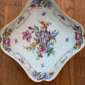 May include: A white porcelain bowl with a diamond-shaped silhouette and scalloped edges. The interior features a central floral bouquet with purple, pink, orange, and blue flowers, surrounded by gold-trimmed scrollwork along the rim.
