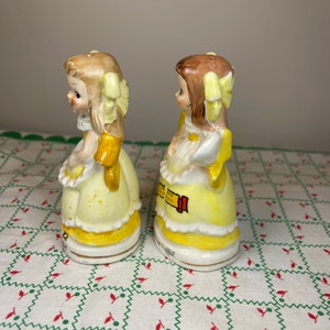 K&ouml;nnte beinhalten: Ein Paar vintage Salz- und Pfefferstreuer mit zwei M&auml;dchen in gelben Kleidern mit gelben Schleifen im Haar. Die Streuer sind aus Keramik und haben ein handbemaltes Design.