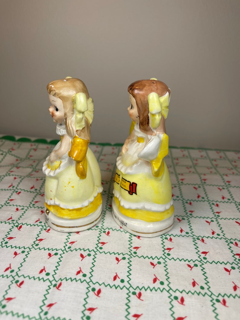 K&ouml;nnte beinhalten: Ein Paar vintage Salz- und Pfefferstreuer mit zwei M&auml;dchen in gelben Kleidern mit gelben Schleifen im Haar. Die Streuer sind aus Keramik und haben ein handbemaltes Design.