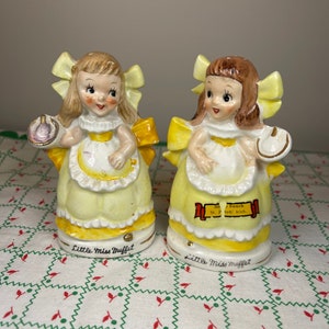 K&ouml;nnte beinhalten: Zwei Porzellanfiguren von Little Miss Muffet, jede mit einer Sch&uuml;ssel in der Hand. Eine Figur tr&auml;gt ein gelbes Kleid mit einer violetten Sch&uuml;ssel, die andere ein gelbes Kleid mit einer wei&szlig;en Sch&uuml;ssel. Beide Figuren haben gelbe Schleifen im Haar und stehen auf einer wei&szlig;en Tischdecke mit rotem und gr&uuml;nem Muster.
