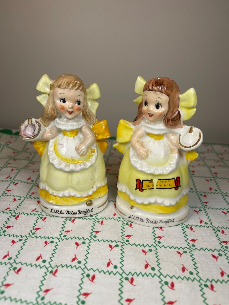 K&ouml;nnte beinhalten: Zwei Porzellanfiguren von Little Miss Muffet, jede mit einer Sch&uuml;ssel in der Hand. Eine Figur tr&auml;gt ein gelbes Kleid mit einer violetten Sch&uuml;ssel, die andere ein gelbes Kleid mit einer wei&szlig;en Sch&uuml;ssel. Beide Figuren haben gelbe Schleifen im Haar und stehen auf einer wei&szlig;en Tischdecke mit rotem und gr&uuml;nem Muster.