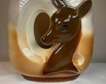 May include: A ceramic planter with a brown deer head design. The planter is glazed in a light brown and white color scheme. The deer head is in relief and has a hole in the center of the planter.