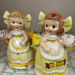 K&ouml;nnte beinhalten: Ein Paar vintage Salz- und Pfefferstreuer mit Little Miss Muffet. Die Streuer sind aus Keramik und in Gelb, Wei&szlig; und Braun bemalt. Die Streuer haben die Form eines M&auml;dchens, das ein gelbes Kleid und eine wei&szlig;e Sch&uuml;rze tr&auml;gt. Die Streuer haben eine gelbe Schleife auf dem Kopf und halten eine Sch&uuml;ssel und einen L&ouml;ffel. Die Streuer sind mit "Little Miss Muffet" und "Silver Beach St. Joseph Mich." beschriftet.