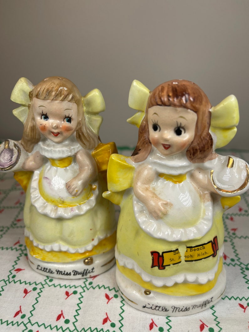K&ouml;nnte beinhalten: Ein Paar vintage Salz- und Pfefferstreuer mit Little Miss Muffet. Die Streuer sind aus Keramik und in Gelb, Wei&szlig; und Braun bemalt. Die Streuer haben die Form eines M&auml;dchens, das ein gelbes Kleid und eine wei&szlig;e Sch&uuml;rze tr&auml;gt. Die Streuer haben eine gelbe Schleife auf dem Kopf und halten eine Sch&uuml;ssel und einen L&ouml;ffel. Die Streuer sind mit "Little Miss Muffet" und "Silver Beach St. Joseph Mich." beschriftet.
