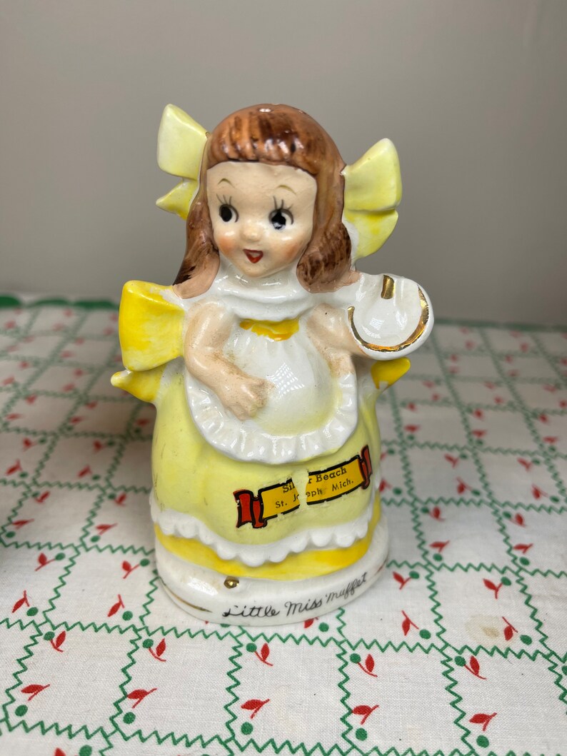 K&ouml;nnte beinhalten: Eine Porzellanfigur eines M&auml;dchens in einem gelben Kleid und einer gelben Schleife im Haar. Sie h&auml;lt ein Hufeisen in der Hand. Die Figur ist mit "Silver Beach St. Joseph Mich." und "Little Miss Muffet" beschriftet.