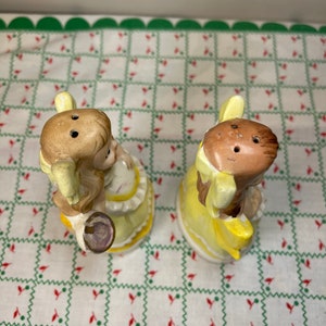 K&ouml;nnte beinhalten: Ein Paar vintage Salz- und Pfefferstreuer in Form von M&auml;dchen in gelben Kleidern. Die Streuer sind aus Keramik und haben ein handbemaltes Design.