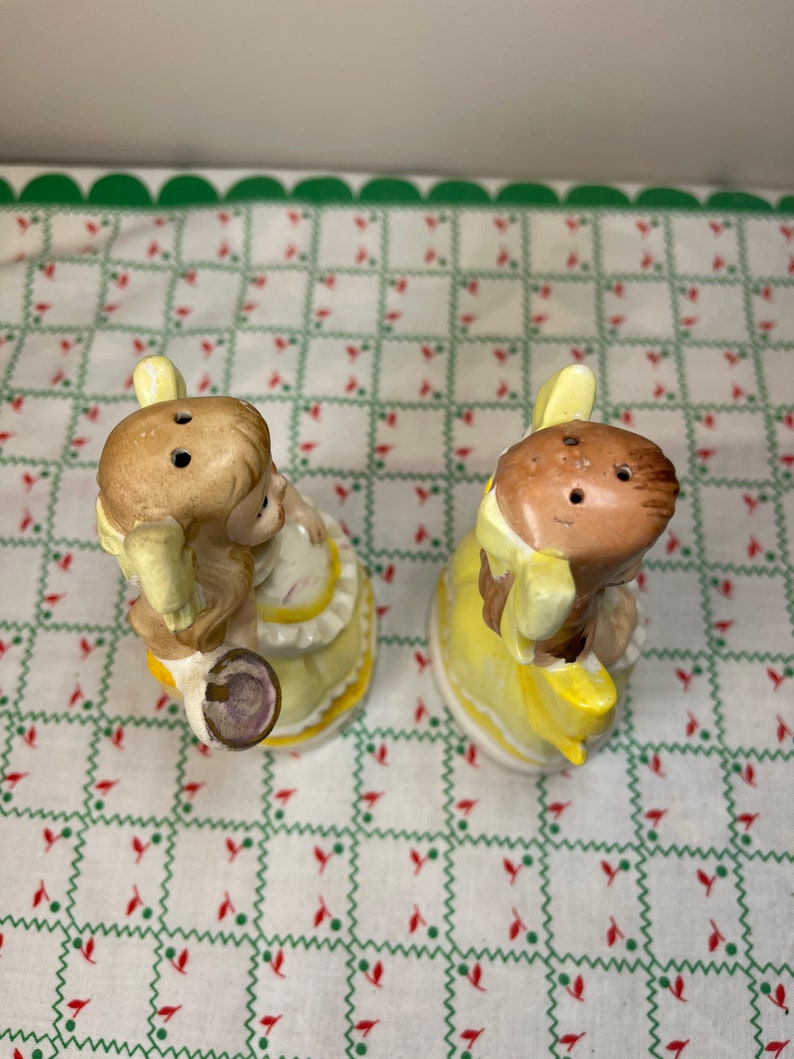 K&ouml;nnte beinhalten: Ein Paar vintage Salz- und Pfefferstreuer in Form von M&auml;dchen in gelben Kleidern. Die Streuer sind aus Keramik und haben ein handbemaltes Design.