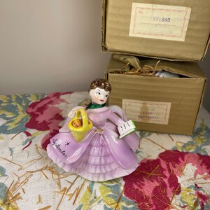 May include: A porcelain figurine of a woman in a purple dress holding a basket of fruit and a book. The figurine is labeled "September" and is part of a collection of figurines representing each month of the year.
