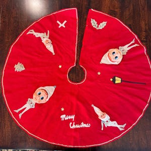 May include: Red felt Christmas tree skirt with a white trim and gold stars. The skirt features appliques of elves, a Christmas tree, a lamppost, and holly. The text "Merry Christmas" is embroidered in white on the skirt.