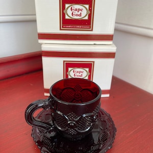 May include: A set of two boxes with the text "Cape Cod" in red and gold lettering. The boxes are stacked on top of each other. A dark red glass teacup and saucer with a decorative pattern is sitting on a red surface.
