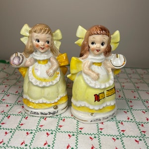 K&ouml;nnte beinhalten: Ein Paar antike Salz- und Pfefferstreuer aus Keramik mit Little Miss Muffet. Die Streuer sind gelb und wei&szlig; mit einer gelben Schleife und einem gelben Kleid. Die Streuer halten eine Sch&uuml;ssel mit Quark und Molke und einen L&ouml;ffel.
